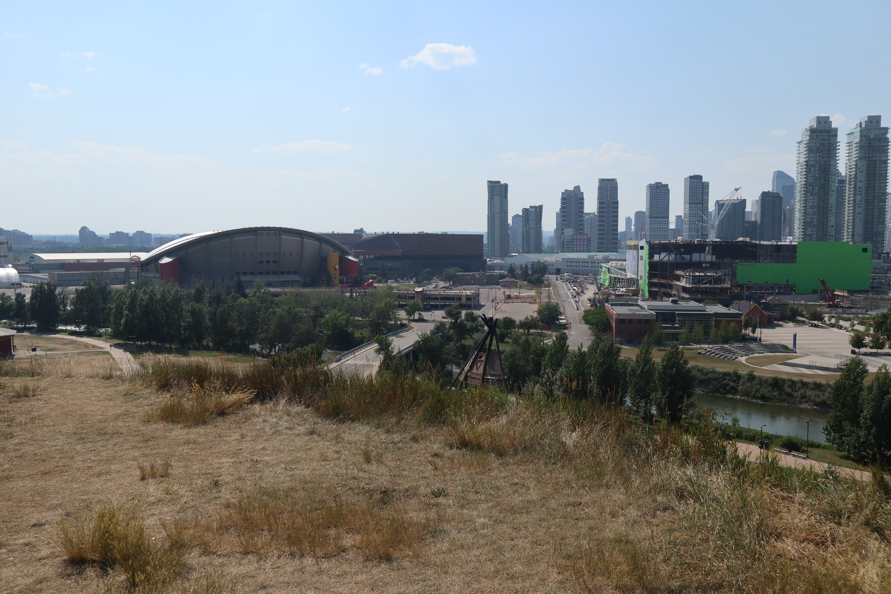 View from Scotsman's Hill looking northwest: the Scotiabank Saddledome's saddle-shaped roof at left, the steel frame of Scotia Place under construction at right, downtown Calgary towers behind, with dry summer grass and a teepee frame in the foreground.