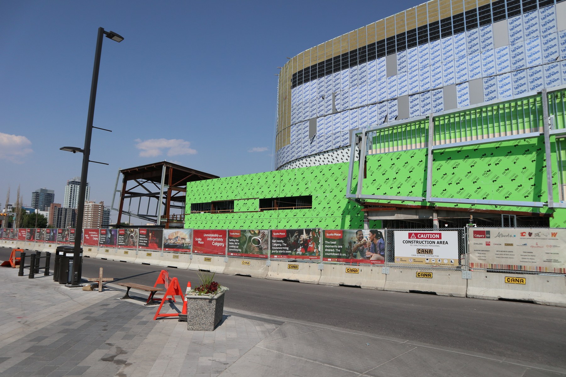Street-level view of the Scotia Place construction site: green weather wrap and insulation panels on the lower levels, white house wrap above, and a long fence line of red Scotia Place marketing banners on concrete barriers stamped with the CANA logo.