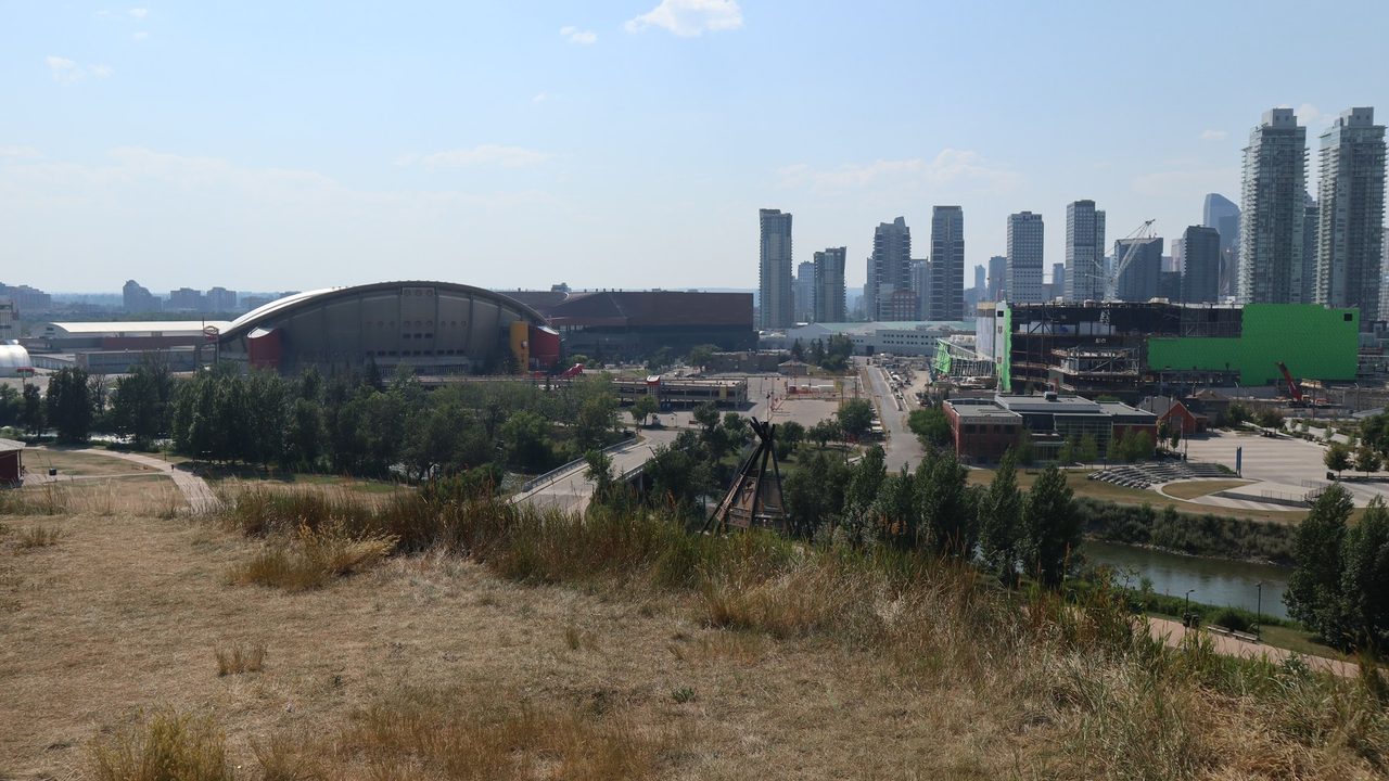 View from Scotsman's Hill looking northwest: the Scotiabank Saddledome's saddle-shaped roof at left, the steel frame of Scotia Place under construction at right, downtown Calgary towers behind, with dry summer grass and a teepee frame in the foreground.