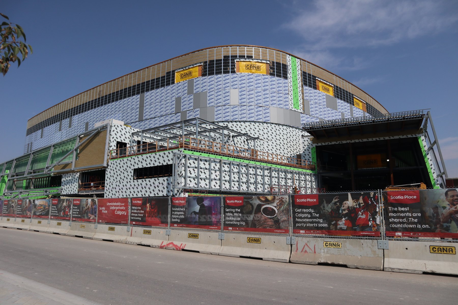The west face of Scotia Place under construction: a continuous tan roof fascia band above white perforated metal panel and printed house wrap, exposed steel and green weather wrap at the left edge where the cladding stops, and a long run of red Scotia Place hoarding banners on CANA-stamped concrete barriers along the bottom.