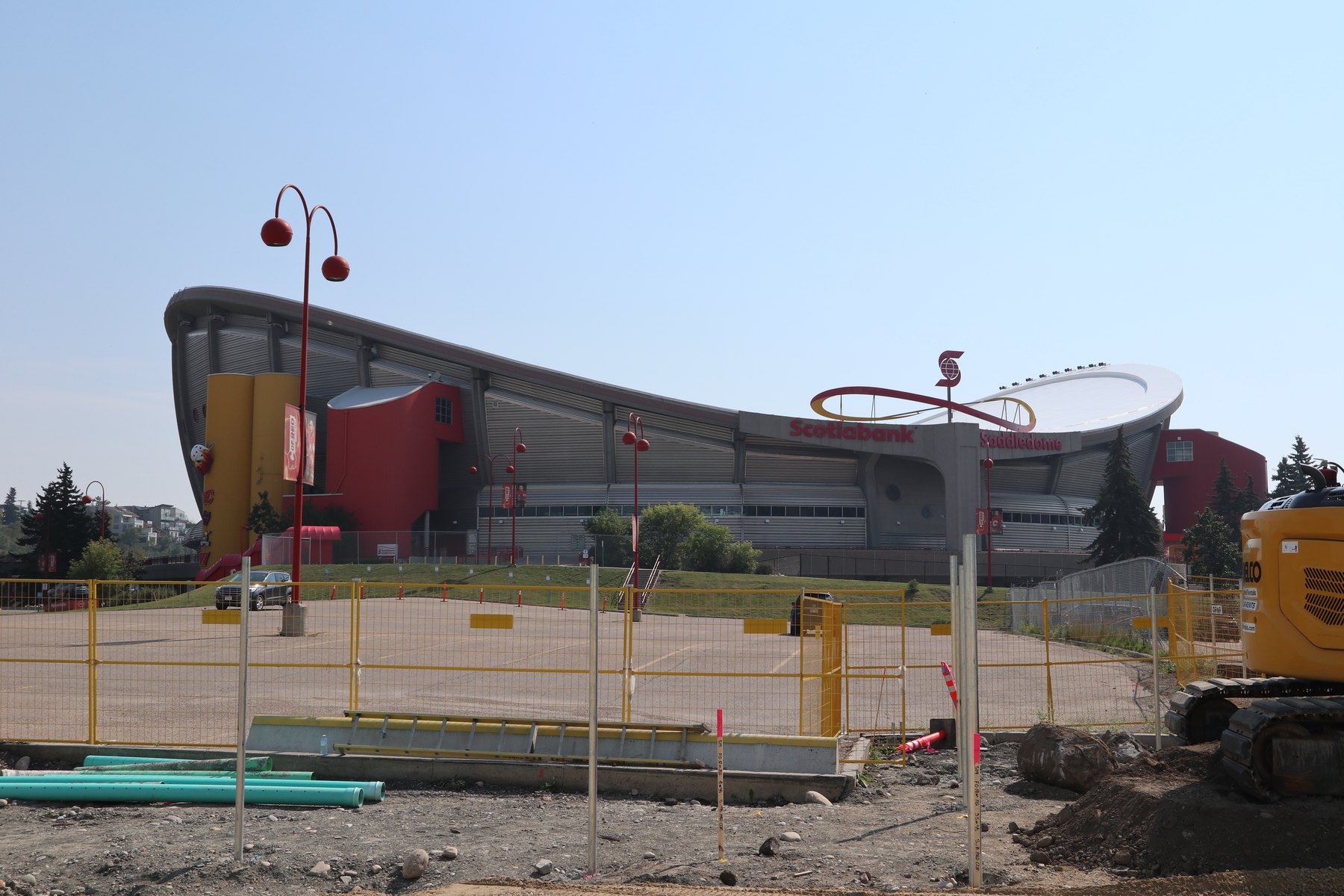 The Scotiabank Saddledome seen from its east parking lot, with an open trench, lengths of green plastic conduit, yellow temporary fencing and a tracked excavator in the foreground.