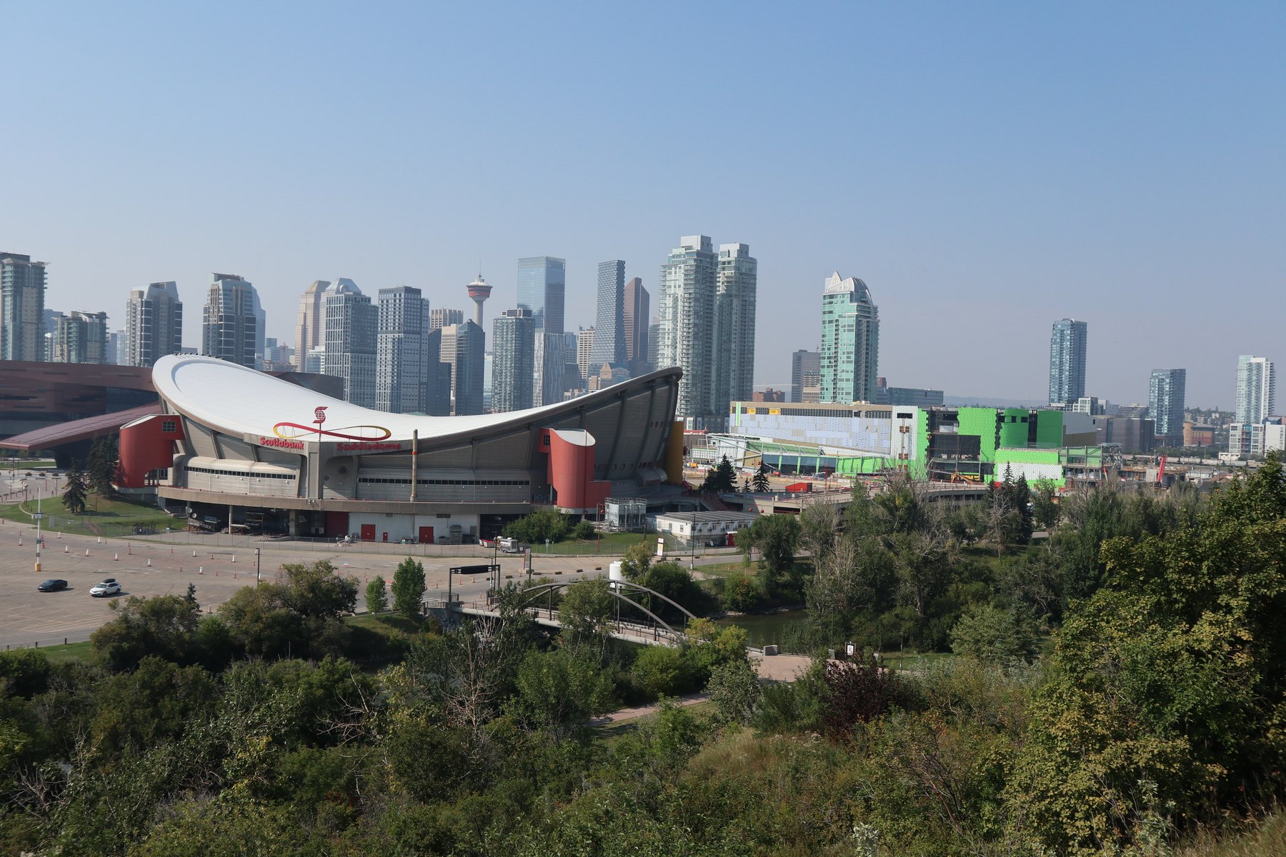 View from Scotsman's Hill: the Scotiabank Saddledome large in the left foreground with downtown Calgary and the Calgary Tower behind it, the Bow River and a pedestrian bridge below, and the green-wrapped east face of Scotia Place at right.