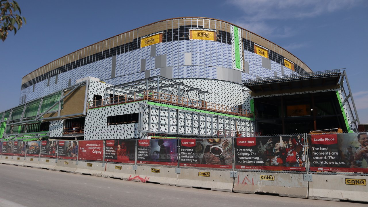 The west face of Scotia Place under construction: a continuous tan roof fascia band above white perforated metal panel and printed house wrap, exposed steel and green weather wrap at the left edge where the cladding stops, and a long run of red Scotia Place hoarding banners on CANA-stamped concrete barriers along the bottom.