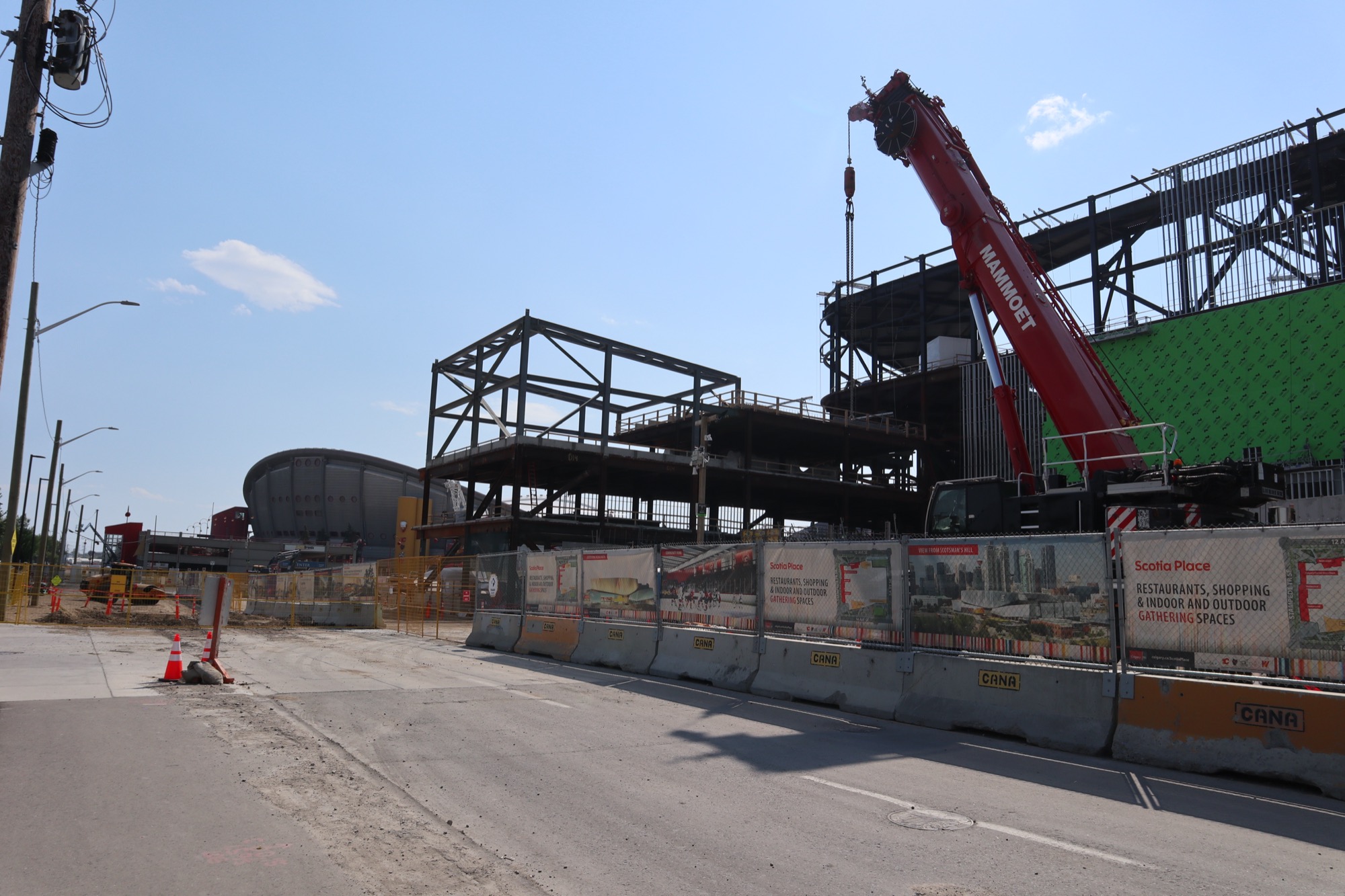 Ground-level view from the street: the Scotiabank Saddledome's curved roof sits low at left, while the steel frame and green-wrapped exterior of Scotia Place rises at right behind a red MAMMOET crane, with CANA-stamped concrete barriers and Scotia Place hoarding running along the sidewalk.