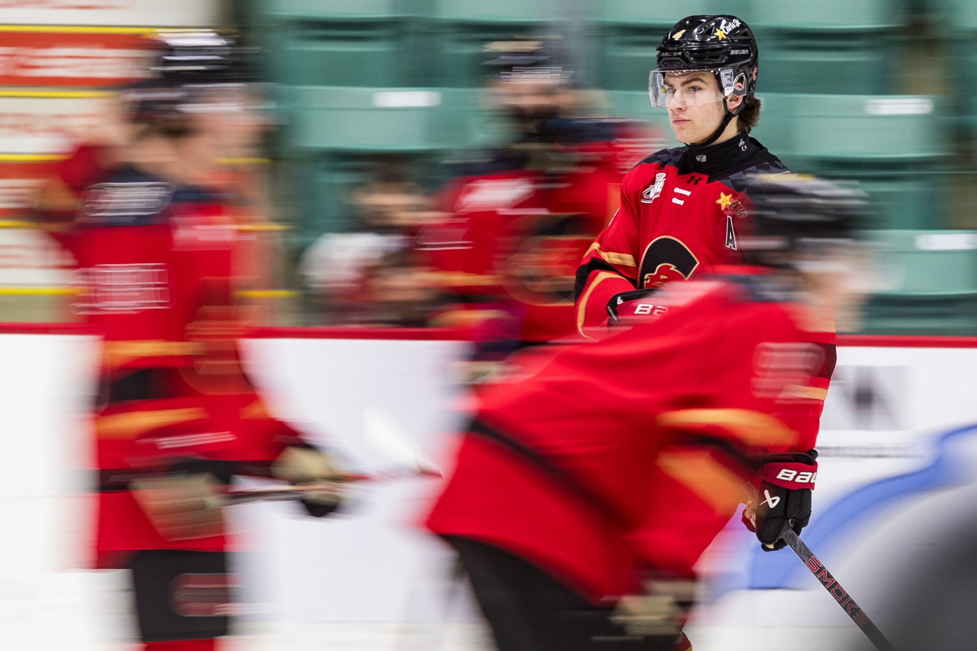Carson Carels in the Prince George Cougars red home jersey with an alternate captain's A, in focus and looking back over his shoulder while teammates streak past in motion blur during a pregame skate.