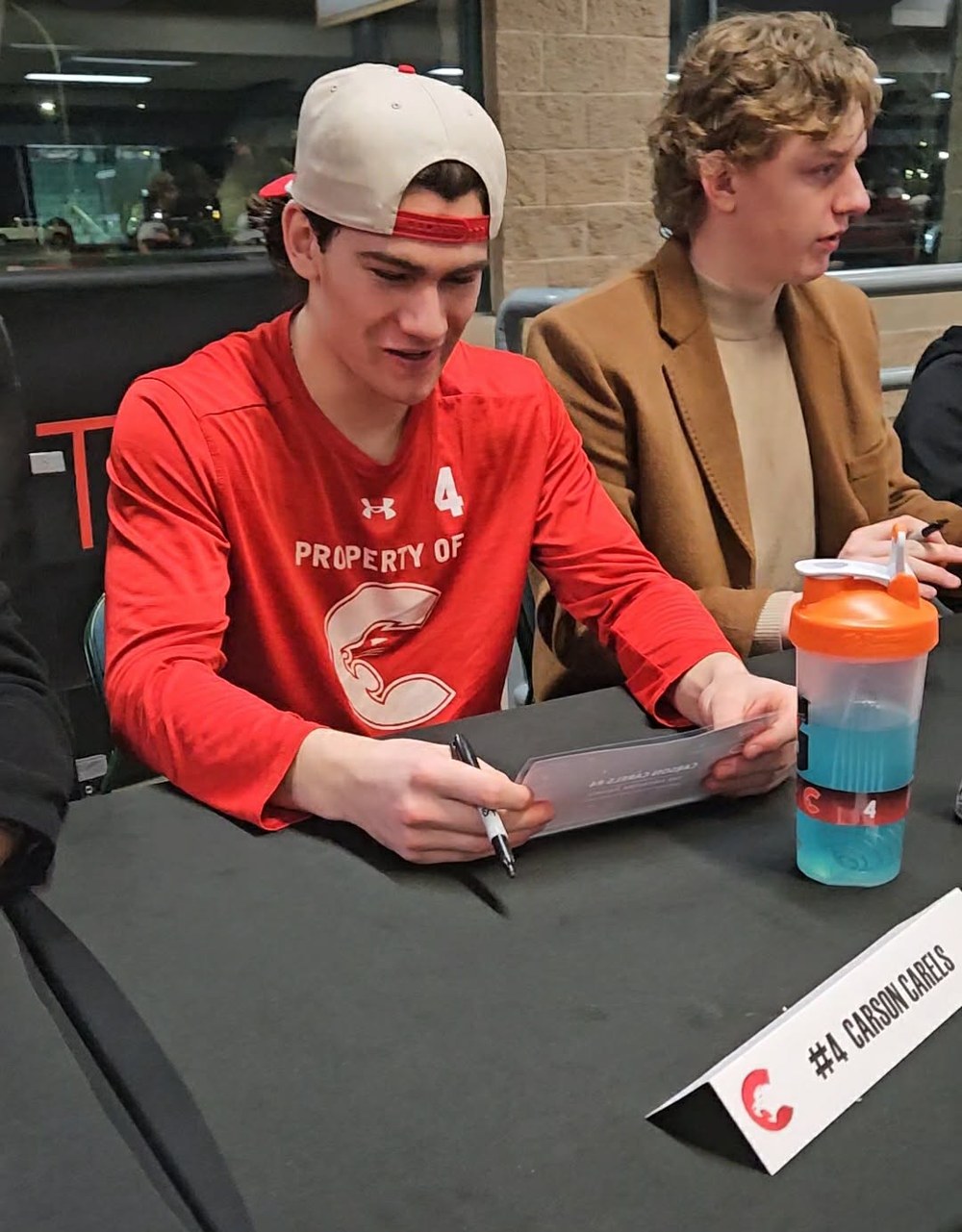 Carson Carels in a red Property of Cougars shirt at a team signing table, reading the Firework Award certificate, his number 4 nameplate in front of him.