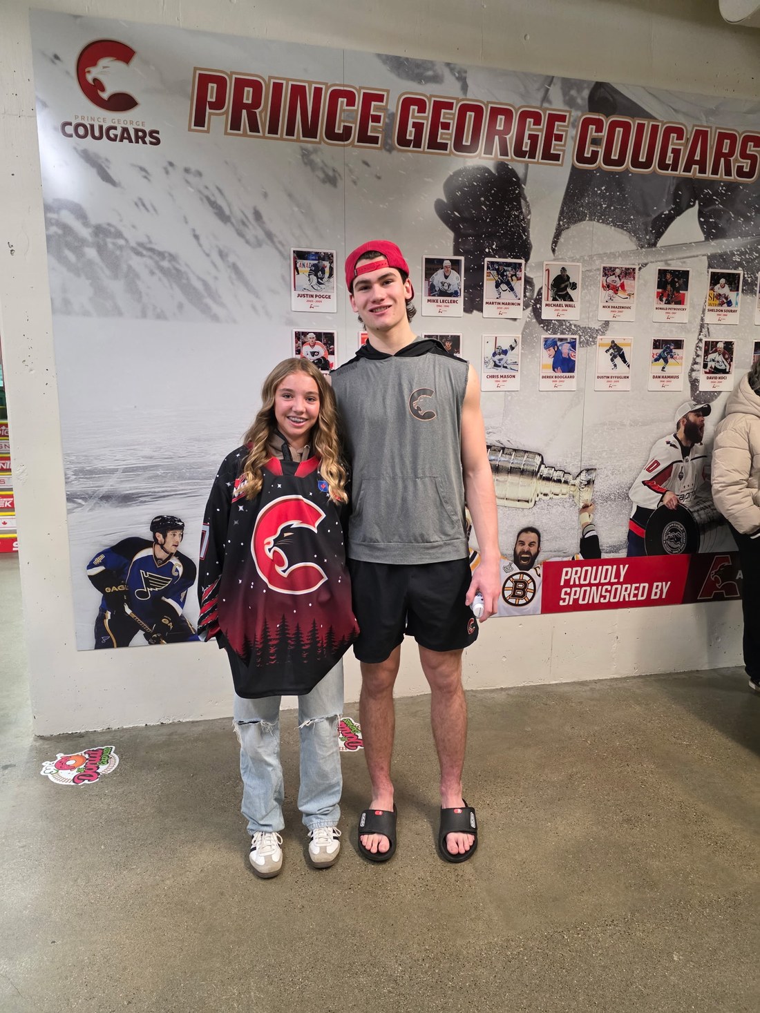 Olivia W. wearing the Cougars jersey she designed, a black sweater with a starry northern sky and treeline, standing with Carson Carels in the CN Centre concourse.