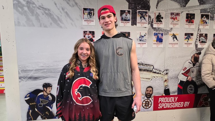 Olivia W. wearing the Cougars jersey she designed, a black sweater with a starry northern sky and treeline, standing with Carson Carels in the CN Centre concourse.